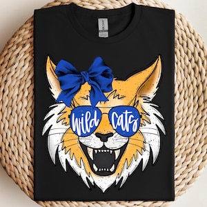 May include: Black t-shirt featuring a cartoon wildcat face with blue sunglasses and a blue bow. The sunglasses have the words "wild cats" in white. The wildcat is orange, white, and black.