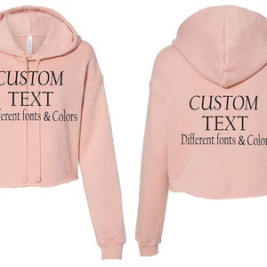 May include: A pink cropped hoodie with a drawstring hood. The front of the hoodie has the text "CUSTOM TEXT Different fonts & Colors" printed on it. The back of the hoodie has the same text printed on it.