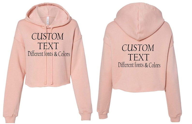 May include: A pink cropped hoodie with a drawstring hood. The front of the hoodie has the text "CUSTOM TEXT Different fonts & Colors" printed on it. The back of the hoodie has the same text printed on it.