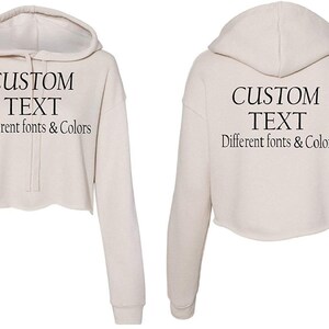 May include: A cream-colored cropped hoodie with a drawstring hood. The hoodie has long sleeves and the text "CUSTOM TEXT Different fonts & Colors" is printed on the front and back.
