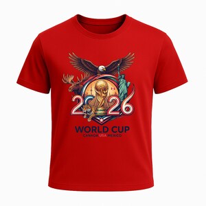 May include: Red t-shirt featuring a graphic for the 2026 World Cup. The design includes an eagle, moose, tiger, Statue of Liberty, and the World Cup trophy. The text "WORLD CUP CANADA USA MEXICO" is printed below the graphic.