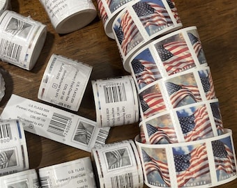 May include: Multiple rolls of U.S. Flags Forever postage stamps featuring a waving American flag design against a blue sky. The rolls include white paper labels with barcodes, the text U.S. FLAGS 2024, and First-Class Rate, manufactured by APU.