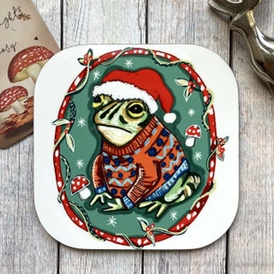 May include: A square coaster featuring a frog wearing a red Santa hat and an orange sweater with blue and white patterns. The frog is inside a green circle with red accents and snowflakes. Small mushroom illustrations are also present.