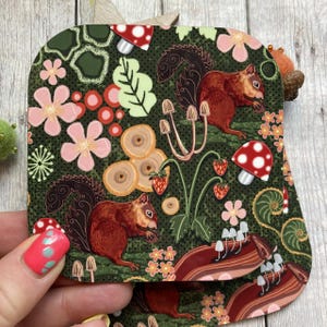 May include: Two brown squirrels with bushy tails are depicted in a whimsical forest scene. The background features a green, leafy pattern with red and pink flowers, mushrooms, and berries.