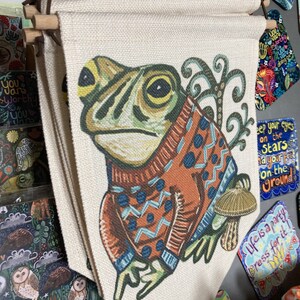 May include: Several canvas wall hangings with a frog illustration. The frog is wearing an orange sweater with blue patterns. The wall hangings have wooden dowels and leather cords at the top. The background is beige.