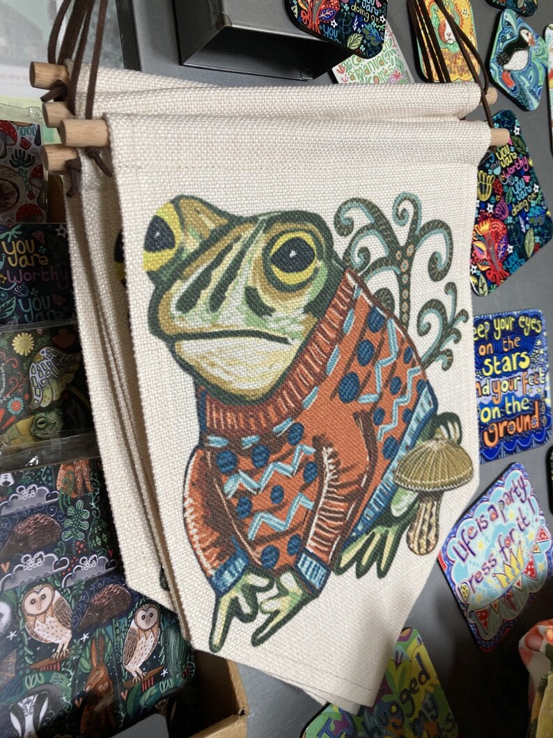 May include: Several canvas wall hangings with a frog illustration. The frog is wearing an orange sweater with blue patterns. The wall hangings have wooden dowels and leather cords at the top. The background is beige.