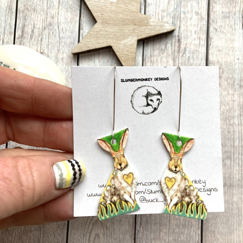 May include: A pair of dangle earrings featuring a green and yellow rabbit design with a heart on its chest. The earrings are on a white card with the text "Slumbermonkey Designs" and a website address.