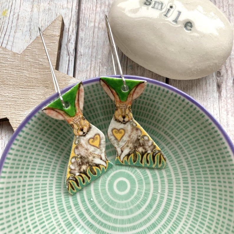 May include: A pair of dangle earrings featuring a green background with a brown rabbit illustration. The rabbit has a yellow heart on its chest and is surrounded by green foliage.