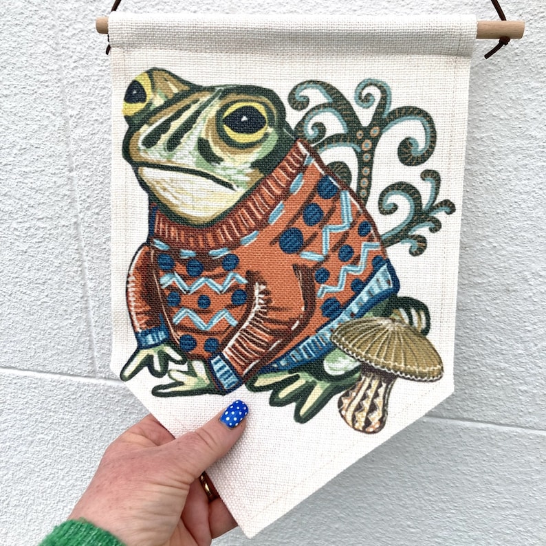 May include: A decorative wall hanging with a frog illustration. The frog is wearing an orange sweater with blue patterns. A mushroom and foliage are also depicted. The banner is held by a wooden rod, with a cream-colored background.