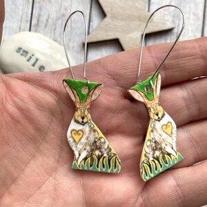 May include: A pair of dangle earrings featuring a whimsical illustration of a rabbit with a heart on its chest. The rabbit is surrounded by green and yellow flames. The earrings are made of metal and have a silver finish.