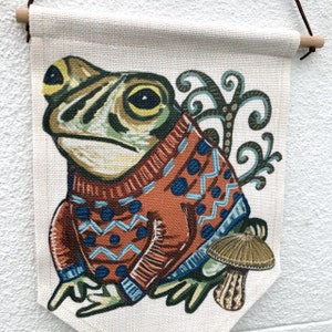 May include: A wall hanging with a frog illustration wearing an orange sweater with blue patterns. The frog is green and yellow, with a mushroom beside it. The banner is made of natural fabric and has a wooden dowel at the top for hanging. The overall design is whimsical.