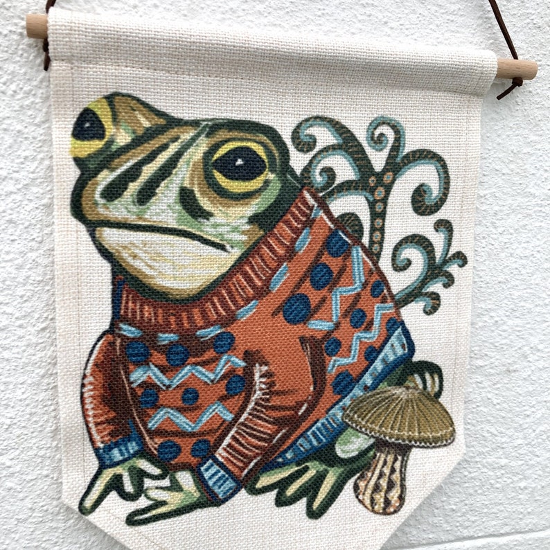 May include: A wall hanging with a frog illustration wearing an orange sweater with blue patterns. The frog is green and yellow, with a mushroom beside it. The banner is made of natural fabric and has a wooden dowel at the top for hanging. The overall design is whimsical.
