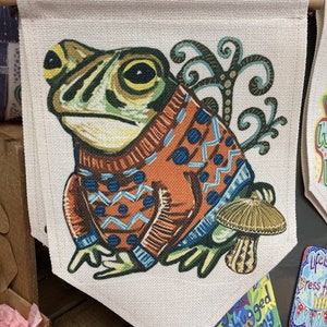 May include: A decorative wall hanging featuring a whimsical frog wearing an orange and blue sweater. The frog is set against a beige background, with a mushroom and decorative swirls. The banner is suspended from a wooden rod.
