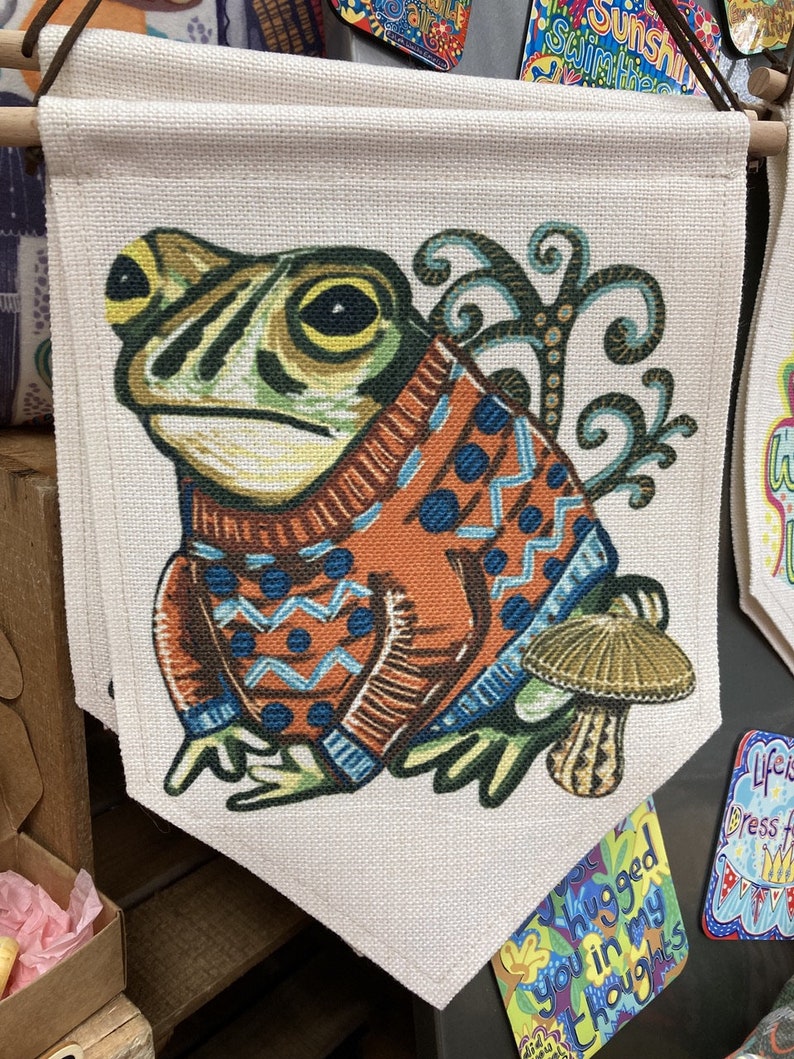 May include: A decorative wall hanging featuring a whimsical frog wearing an orange and blue sweater. The frog is set against a beige background, with a mushroom and decorative swirls. The banner is suspended from a wooden rod.