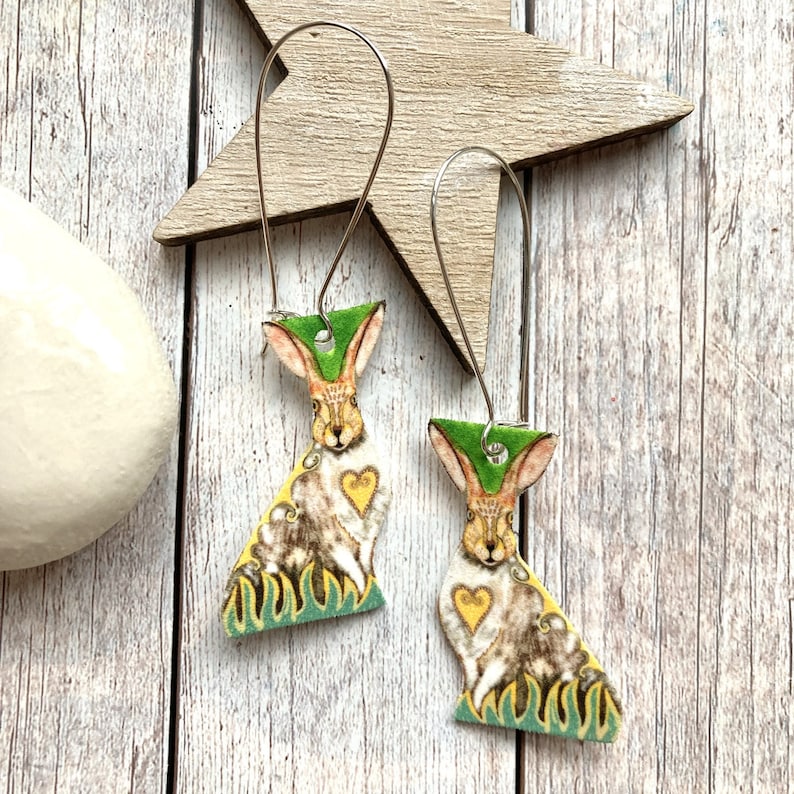 May include: A pair of dangle earrings featuring a whimsical rabbit design. The rabbit is depicted in a brown and white pattern with a yellow heart on its chest. The rabbit is surrounded by green foliage. The earrings are made of metal and have a silver finish.
