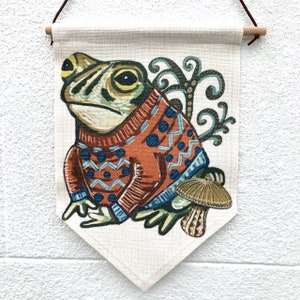 May include: A decorative wall hanging with a frog illustration wearing an orange sweater with blue patterns. The frog is set against a cream background, with a mushroom and decorative swirls. The banner is suspended by a wooden dowel, with a brown cord.