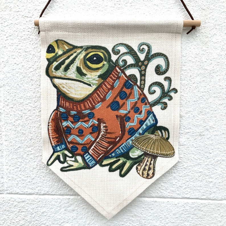 May include: A decorative wall hanging with a frog illustration wearing an orange sweater with blue patterns. The frog is set against a cream background, with a mushroom and decorative swirls. The banner is suspended by a wooden dowel, with a brown cord.