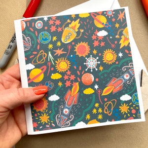 May include: A card with a dark blue background and a colorful pattern of rockets, stars, planets, and suns. The card is perfect for a birthday or other special occasion.