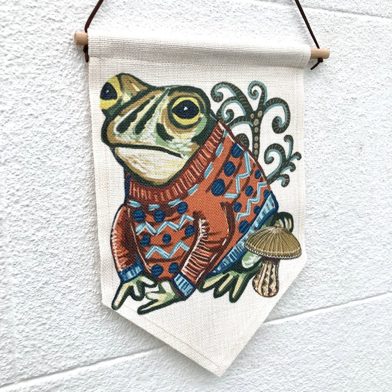 May include: A cream-coloured wall hanging with a frog illustration. The frog is wearing an orange sweater with blue geometric patterns. The artwork includes a mushroom and decorative swirls. The banner is suspended by a wooden dowel and a brown hanging cord.