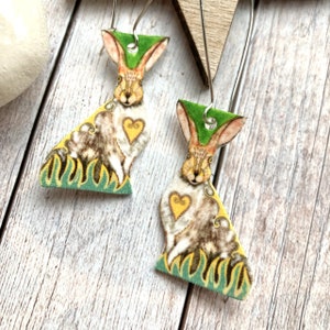 May include: A pair of dangle earrings featuring a whimsical illustration of a brown rabbit with a yellow heart on a green and yellow background.