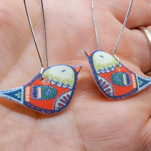 May include: A pair of dangle earrings featuring a colourful bird design. The birds are orange, blue, green and white with intricate patterns. The earrings are made with silver hooks.