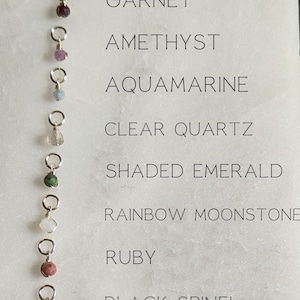 May include: A white card with the names of different gemstones and their corresponding beads. The beads are arranged vertically on the left side of the card, with the names of the gemstones listed on the right side. The beads are all different colors and shapes, and they are attached to silver jump rings.