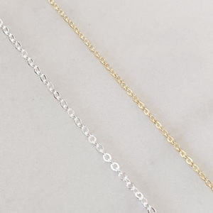 May include: Two chains, one silver and one gold, are laid out on a white surface. The chains are both delicate and have a simple design.