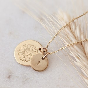 May include: A gold-colored necklace with two pendants. One pendant is a large, round disc with a daisy design etched into it. The other is a smaller, round disc with the letter "T" engraved. The necklace is displayed on a white marble surface.