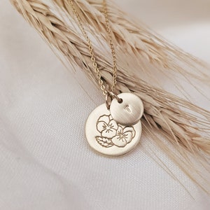 May include: A gold necklace with two charms. One charm is a small circle with the letter "T" engraved on it. The other charm is a larger circle with an engraved design of three pansies.