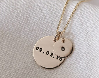 May include: A gold-toned necklace featuring two circular pendants attached to a delicate chain. The larger pendant is stamped with the date 09.03.16, while the smaller pendant is stamped with the letter B. Both pendants have a smooth finish.