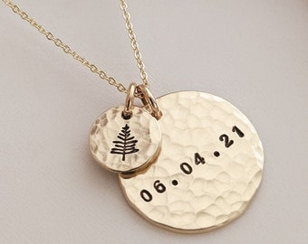May include: A gold-toned necklace featuring two circular, hammered-texture pendants. The smaller pendant displays a black engraved pine tree icon, while the larger pendant is stamped with the black text 06.04.21. Both charms hang from a delicate chain.