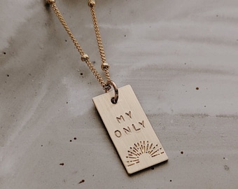 May include: A gold-toned necklace featuring a delicate chain with small beads and a rectangular pendant. The pendant is engraved with the text MY ONLY and a small sunburst graphic at the bottom. The metal has a brushed finish and a simple design.