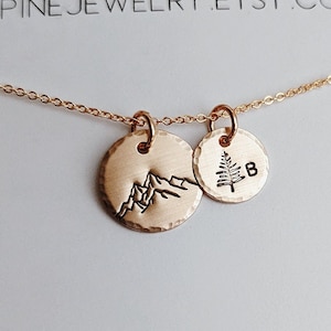 May include: A gold necklace with two charms, one with a mountain silhouette and the other with a pine tree and the letter "B". The necklace is displayed in a white box with the text "wildpinejewelry.etsy.com" and a design of pine trees and stars.