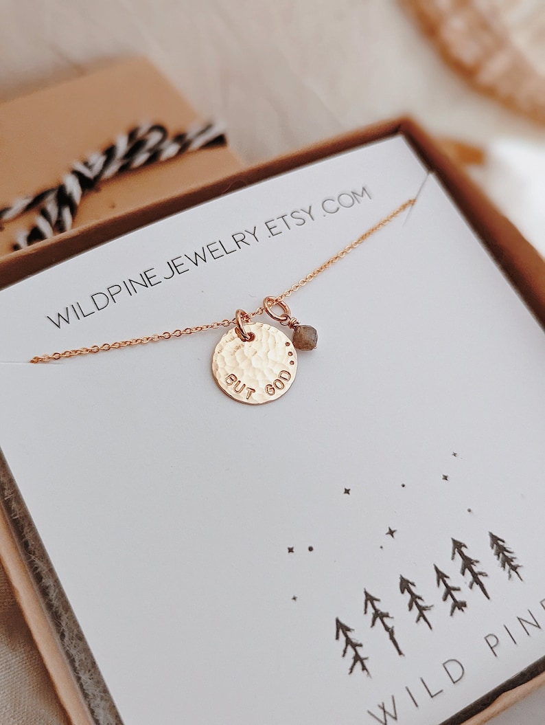 May include: A gold necklace with a round pendant that says "but god" and a small grey gemstone charm. The necklace is in a white box with a black and white striped ribbon and the text "wildpinejewelry.etsy.com" printed on the box.