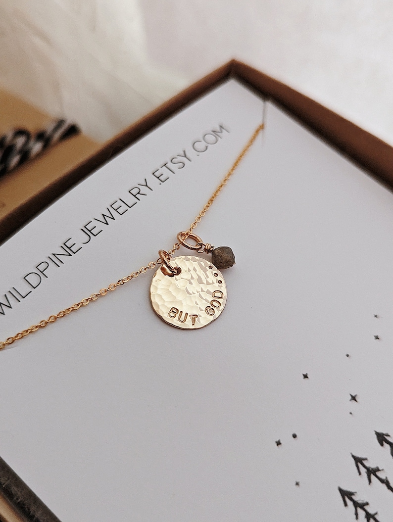 May include: Gold necklace with a hammered disc pendant that says "But God" and a small green gemstone charm. The necklace is in a white box with the text "Wildpine Jewelry Etsy.com" printed on it.