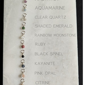 May include: A chart with 12 different gemstone beads on silver jump rings. The chart lists the names of the gemstones from top to bottom: Garnet, Amethyst, Aquamarine, Clear Quartz, Shaded Emerald, Rainbow Moonstone, Ruby, Black Spinel, Kyanite, Pink Opal, Citrine, Green Turquoise.