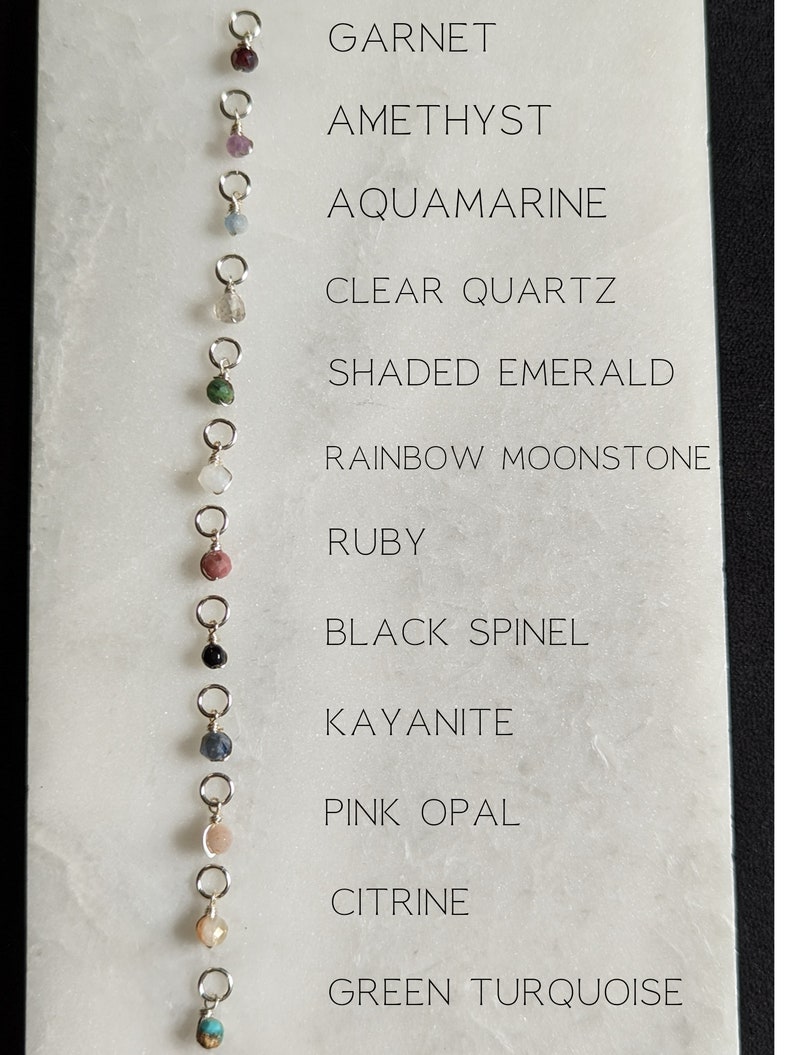 May include: A chart with 12 different gemstone beads on silver jump rings. The chart lists the names of the gemstones from top to bottom: Garnet, Amethyst, Aquamarine, Clear Quartz, Shaded Emerald, Rainbow Moonstone, Ruby, Black Spinel, Kyanite, Pink Opal, Citrine, Green Turquoise.