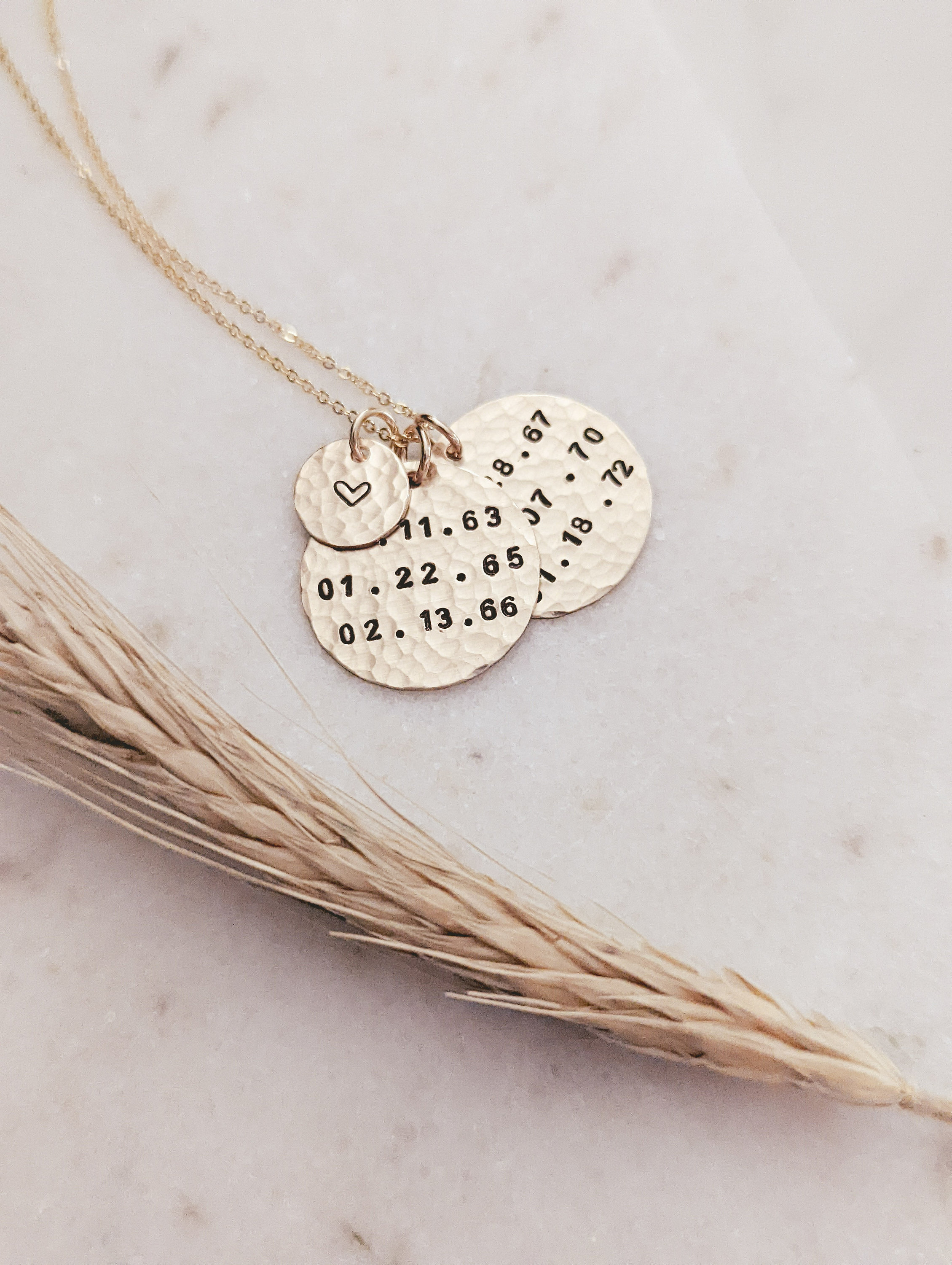 May include: A gold-toned necklace featuring three circular, hammered-texture pendants. The smallest charm displays a heart symbol, while the two larger discs are stamped with dates including 11.63, 01.22.65, 02.13.66, 08.67, 07.70, and 01.18.72 in black ink.