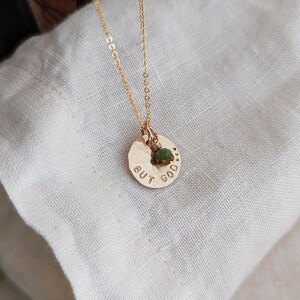 May include: Gold necklace with a hammered disc pendant that says "BUT GOOD." A small green gemstone is attached to the pendant.