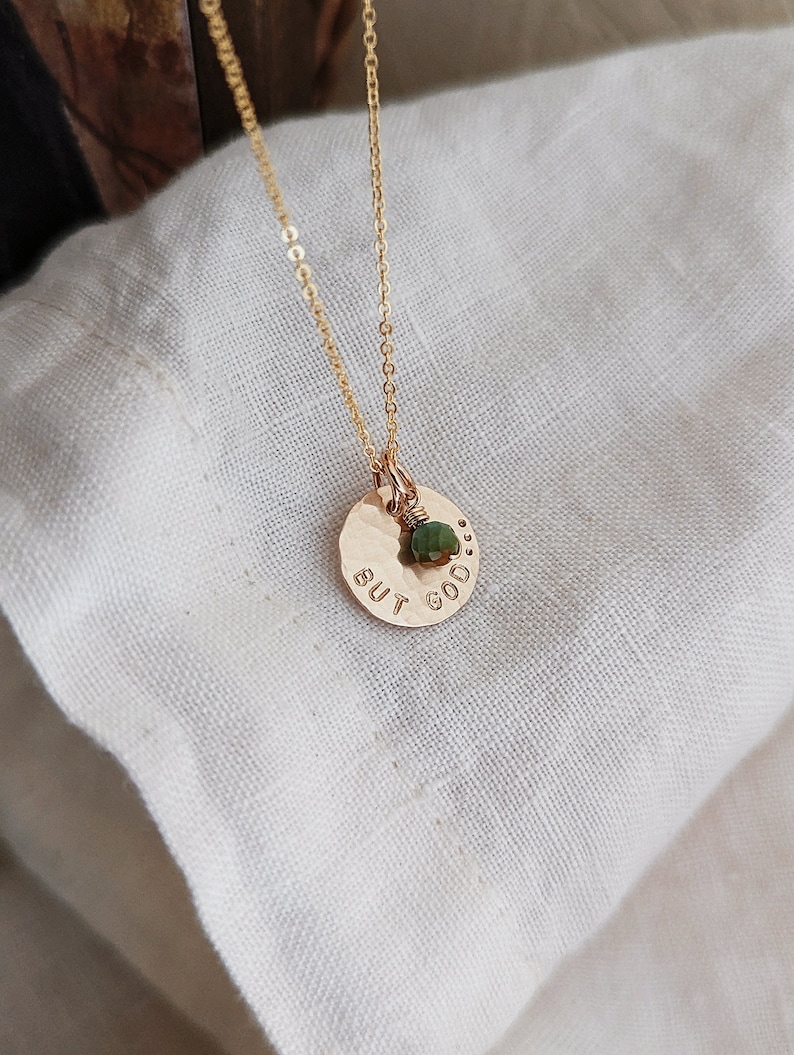 May include: Gold necklace with a hammered disc pendant that says "BUT GOOD." A small green gemstone is attached to the pendant.