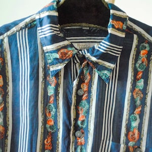 以下が含まれることがあります： 青と白のストライプのボタンダウンシャツ。シャツは、オレンジ、緑、金色の花柄と貝殻柄の装飾的な垂直パネルが特徴です。襟にも同様の模様があります。「Henry Morell」と書かれたラベルが付いています。