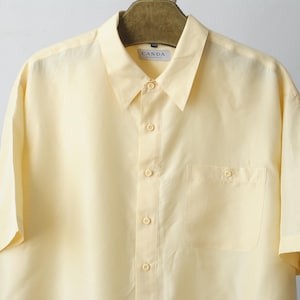 以下が含まれることがあります： ボタンダウンと胸ポケットが付いた、淡いイエローの半袖シャツ。 シャツはクラシックな襟で、木製のハンガーに掛けられています。 ブランド名「CANDA」がラベルに表示されています。