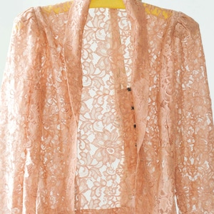 May include: A peach-coloured lace jacket with a floral pattern. The jacket has long sleeves and a shawl collar. It is hanging on a yellow hanger. The delicate lace fabric is visible.
