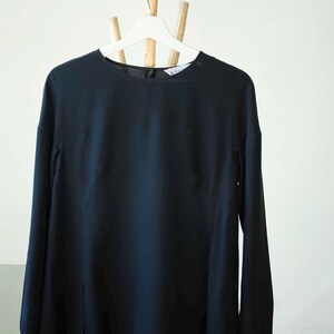 以下が含まれることがあります： 白いハンガーに掛けられた黒い長袖ブラウス。 ブラウスは丸いネックラインとスリット裾が特徴です。 袖口はボタン留めです。 ブラウスは滑らかで流れるような生地で作られています。