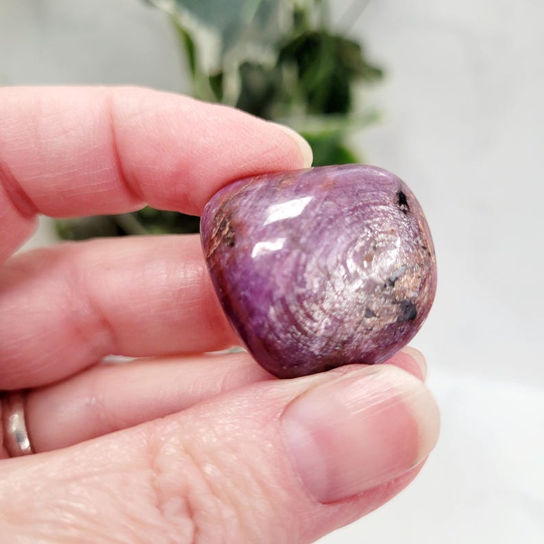 Puede incluir: Una piedra preciosa pulida en forma de coraz&oacute;n, en tonos morados y plateados. La piedra tiene un patr&oacute;n arremolinado y una forma suave y redondeada. La piedra preciosa se sostiene en una mano, con un fondo borroso de vegetaci&oacute;n.