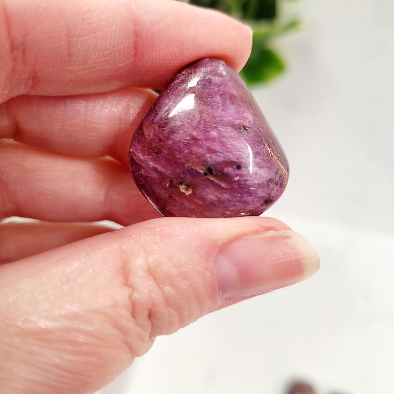 Puede incluir: Una gema pulida en forma de l&aacute;grima, en tonos de p&uacute;rpura intenso y magenta, con sutiles vetas negras y doradas. La piedra se sostiene entre los dedos, mostrando su superficie lisa y reflectante. Esta piedra natural es una pieza &uacute;nica.
