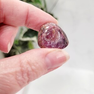 Puede incluir: Una piedra preciosa pulida y rodada en tonos de rosa, morado y marr&oacute;n. La piedra se sostiene entre dos dedos, mostrando su forma suave y redondeada y sus variaciones de color naturales. La piedra preciosa parece transl&uacute;cida.