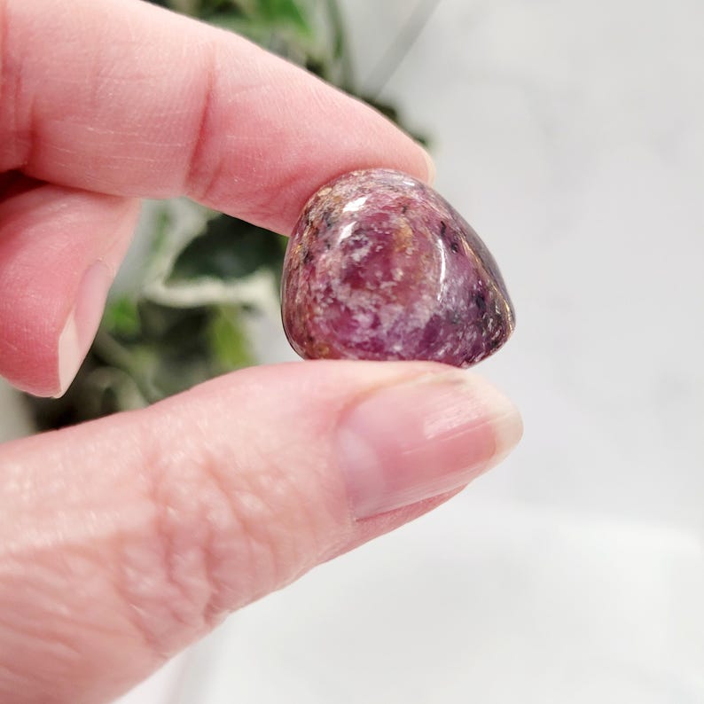 Puede incluir: Una piedra preciosa pulida y rodada en tonos de rosa, morado y marr&oacute;n. La piedra se sostiene entre dos dedos, mostrando su forma suave y redondeada y sus variaciones de color naturales. La piedra preciosa parece transl&uacute;cida.