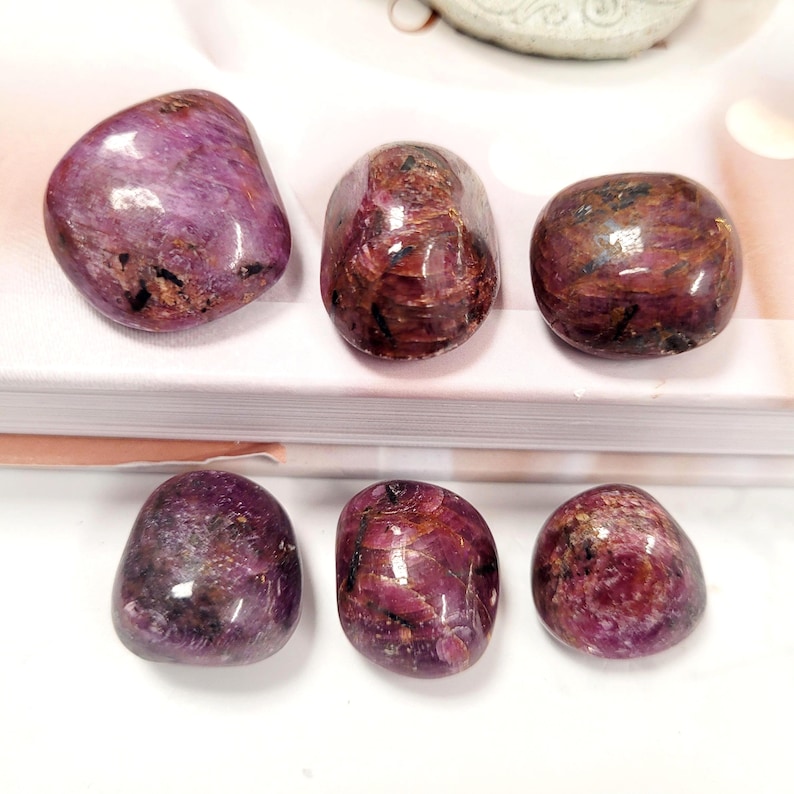 Puede incluir: Seis piedras pulidas y rodadas en tonos rojo intenso y morado. Las piedras tienen un acabado liso y brillante y patrones variables de color y textura. La disposici&oacute;n est&aacute; sobre una superficie blanca.