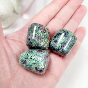 May include: Three polished ruby in zoisite stones, each with a unique pattern of green and red hues. The stones are held in an open hand, showcasing their natural beauty and texture. The stones are smooth and rounded.